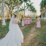 A bride in a white wedding gown approaches her bridesmaids on a tree-lined pathway outdoors.