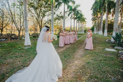 A bride in a white wedding gown approaches her bridesmaids on a tree-lined pathway outdoors.
